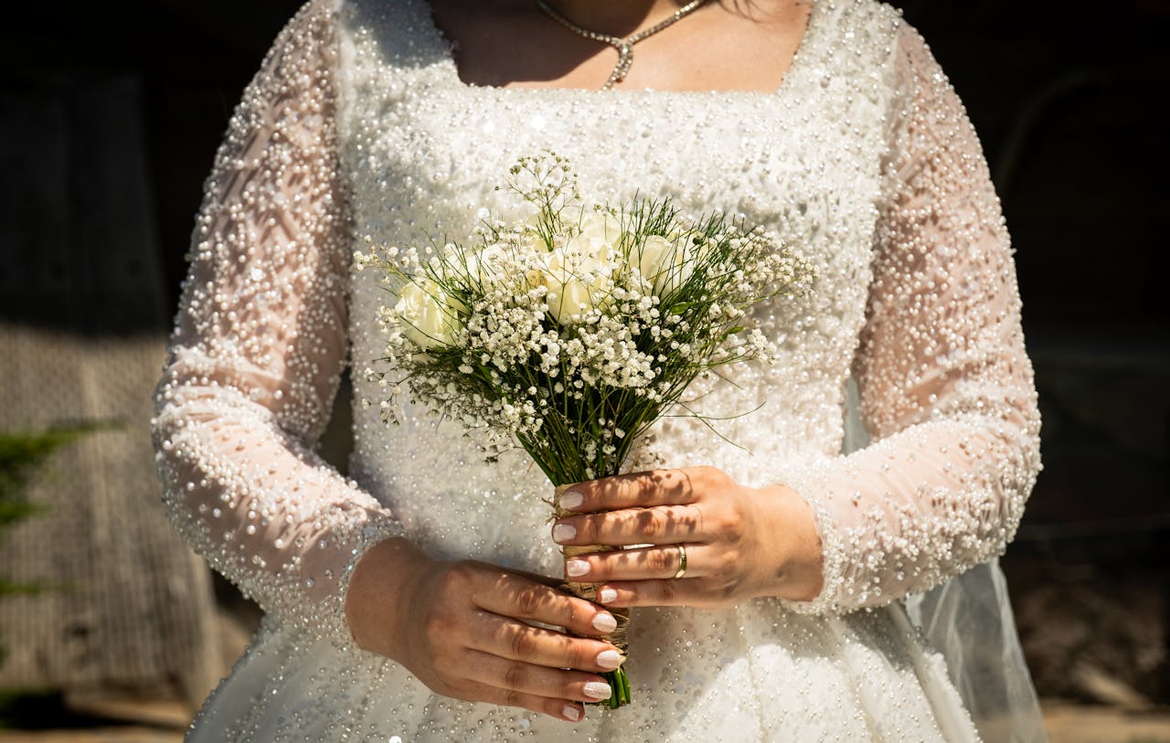 Bride in an embellished wedding dress holding a delicate white bouquet outdoors.