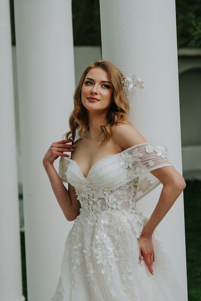 A beautiful bride in a white lace wedding dress poses gracefully against a column outdoors.